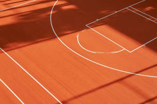 High angle view of bright orange basketball court surface featuring crisp white three point arc and key lines with dramatic geometric shadows cast by overhead sunlight across the textured synthetic