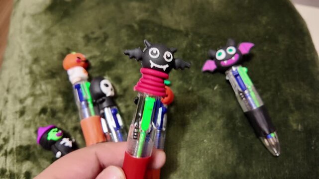 Hand holds Halloween-themed pens against a green backdrop. Pens feature bats, pumpkins, and spooky faces. Each pen has a colorful, playful design. Ideal for festive, creative, or educational uses