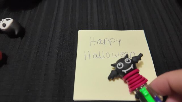Hand writes "Happy Halloween" on sticky note. Black bat charm sits atop note. Green pencil held near note. Dark table holds small Halloween toys. Childlike joy shown in handwritten note