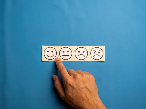 A close up of a human hand with one finger pointing at a smiling face icon on a white paper strip containing four different facial expressions against a solid blue background