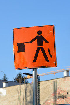 A square orange traffic sign with a black flagman symbol stands on a metal pole near a construction site under a clear blue sky.n with a flagman icon against a blue sky