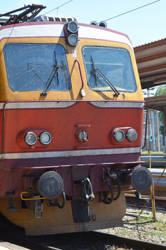 Close up front view of a red and yellow electric passenger train locomotive parked on the tracks under a bright sky.