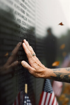 Senior male veteran of war rests against a black memorial wall, his hand tracing a name in a moment of deep, silent grief. Poignant scene of the lasting weight of loss and enduring power of memory