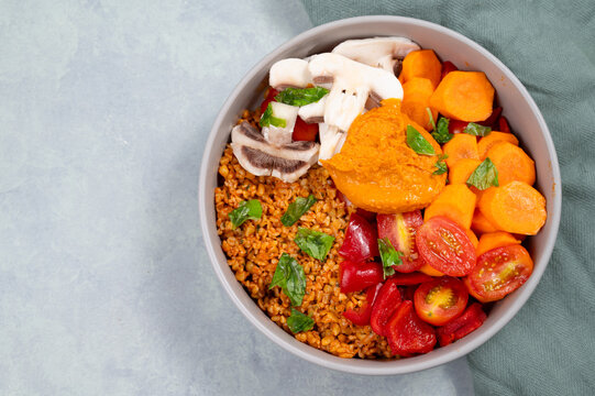 Bowl with bulgur, carrots, cherry tomatoes, red pepper, champignons, hummus dip and fresh basil herbs, healthy vegan food, plant based diet