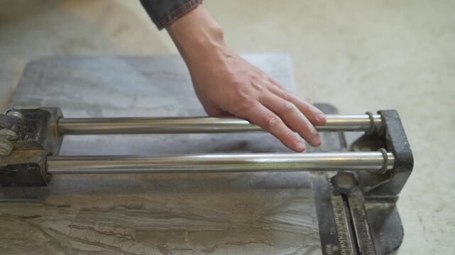 Close up of a construction workers hands scoring a grey marbled floor tile with a manual tile cutter. A detailed shot of home improvement, DIY, or professional tiling work in progress