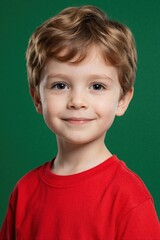 Fototapeta premium Cute little boy with blond hair wears red t-shirt. Smiling young kid looks at camera in studio. Portrait of happy child with bright eyes on green background. Childhood happiness, innocence concept