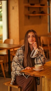Young woman sitting at a wooden table in a cozy cafe with a warm cup of coffee in the afternoon