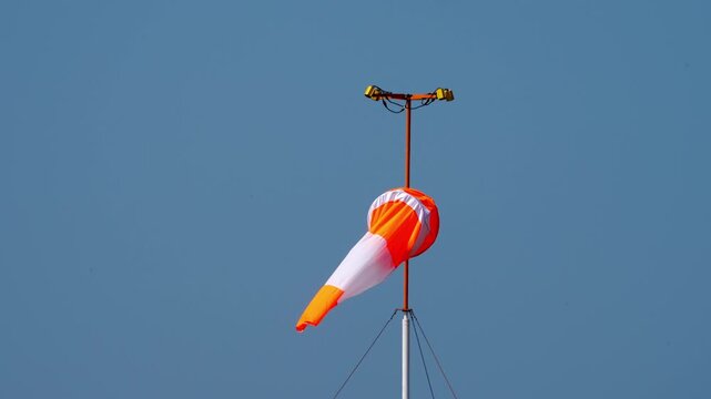 Orange and white striped windsock fluttering in wind on mast with obstruction lights against clear blue sky, wind direction indicator at airfield, close-up