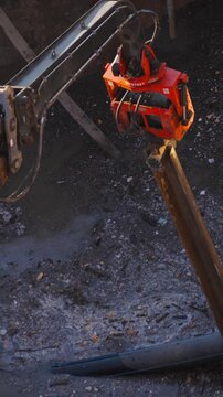 Excavator-mounted vibratory pile driver drives steel sheet pile into pit filled with black foul sewage sludge during emergency sewer repair, vertical video, top view