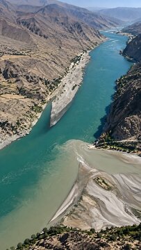 Dramatic aerial shot of a turquoise river confluence merging with a grey sediment stream in a rugged mountain valley, showing textured rocky terrain and winding water paths in an arid environment.