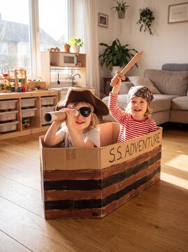 Two young children dressed as pirates, one with an eye patch and the other wearing a bandana, joyfully sail their cardboard box ship named S.S. Adventure through imaginary waters in a sunlit room.