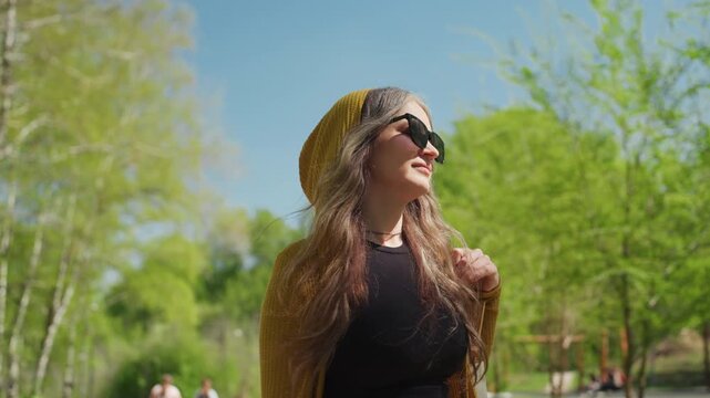 Woman with long hair wears yellow hooded sweater and sunglasses standing outdoors in bright sunlight holding wooden chair parts while looking around. Woman is surrounded by green trees