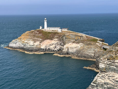 Anglesey, Wales: A white lighthouse and outbuildings stand on top of a rocky island with layered cliffs, surrounded by deep blue sea, with a footbridge and walled path leading to the island.