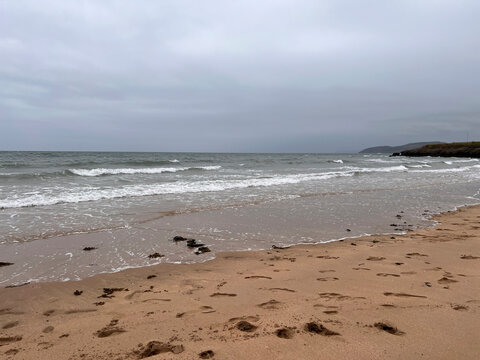 Anglesey, Wales: Waves break and wash up a sandy beach under an overcast grey sky, with footprints and scattered seaweed on the wet sand and a rocky headland on the horizon at right.