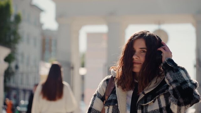 Young woman with wavy hair wears black and white checkered coat and large over ear headphones while walking on city street. Woman carries backpack adjusts her headwear as woman moves