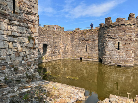 Beaumaris Castle, Isle of Anglesey, - july 17, 2026: Stone curtain walls and a round tower with crenellations rise above a moat of murky green water, as two visitors stand on the wall walk behind a ra