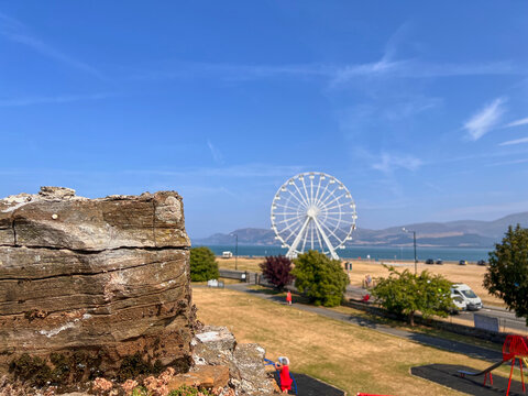Beaumaris Castle, Isle of Anglesey, - july 17, 2026: A weathered stone wall section stands in the foreground, with a white Ferris wheel, a grassy park, a playground and a coastal strait with hills bey