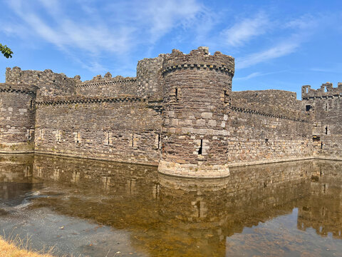 Beaumaris Castle, Isle of Anglesey, - july 17, 2026: Stone curtain walls and a round tower of a medieval castle rise above a still moat that reflects the masonry under a blue sky with thin clouds