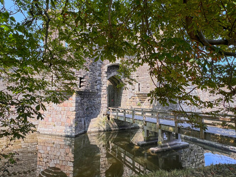 Beaumaris Castle, Isle of Anglesey, - july 17, 2026: A wooden footbridge crosses a moat to a stone gatehouse with an arched entrance and iron gate, framed by horse chestnut branches and reflected in t