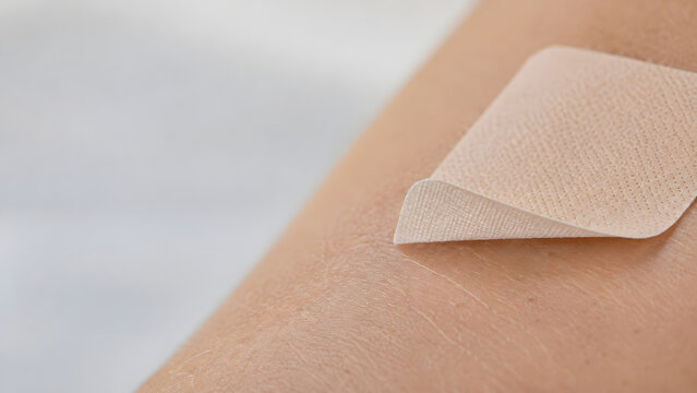 Adhesive pain relief patch is partially lifted from bare skin in a macro view, revealing a thin flexible fabric surface, a raised corner and natural skin texture against a soft neutral background.