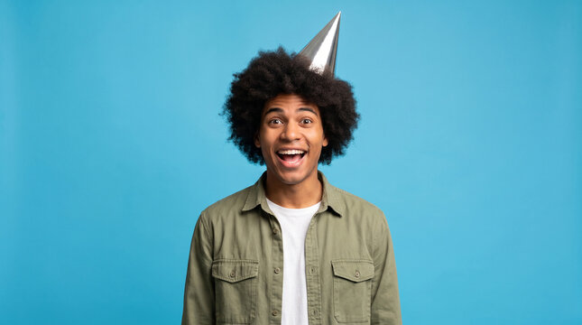 Excited young man with an afro hairstyle wearing a party hat and a green shirt, celebrating International Men's Day with a joyful expression.