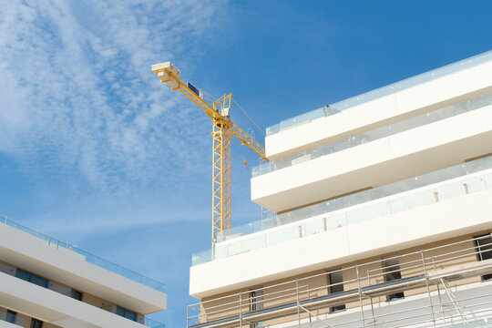 Low angle view of tower crane and modern white apartment buildings under construction against blue sky
