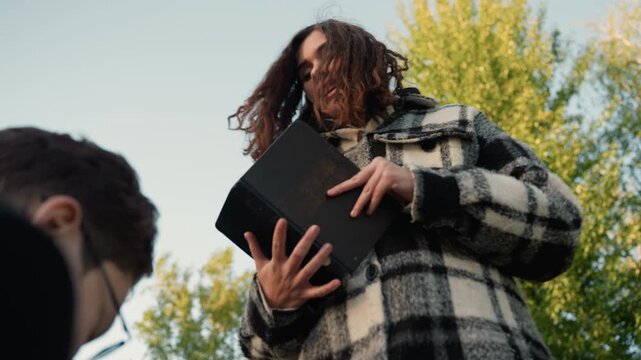 Woman stands outdoors holding open book while another woman is near. Woman wears plaid jacket and focuses on text as others are present in background outdoor