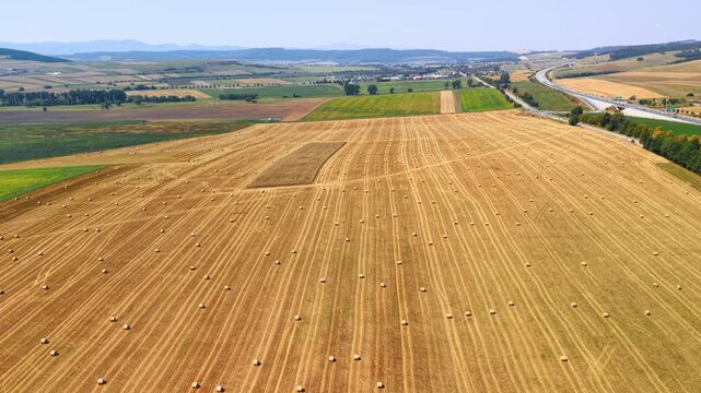 Harvest rows across a field beside a highway in Slovakia. Numerous round bales sit on cut farmland, with patchwork crops, a highway and hills beyond.