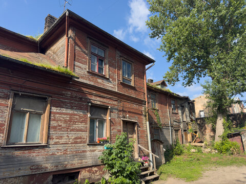 Old wooden residential building in Riga, Latvia, with peeling red paint, weathered timber siding and aging windows. Overgrown vegetation and a rustic courtyard surround the deteriorating structure on 