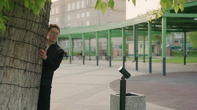 Man dressed entirely in black partially hides against rough bark of large urban tree. background reveals open public area with green metal structures distant buildings and soft natural light