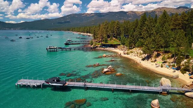 Piers along a forested Lake Tahoe cove. Wooden docks extend over turquoise water beside a sandy beach, boulders and conifers below mountains.