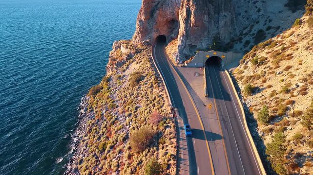 Twin road tunnels beside Lake Tahoe. Vehicles approach two cliff portals above blue water, with guardrails and conifers along the slope.