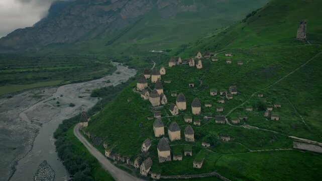 Aerial footage shows the medieval stone crypts of the ancient necropolis in Dargavs, North Ossetia-Alania, Russia, situated on a green mountain hill near a riverbed under dramatic cloudy skies
