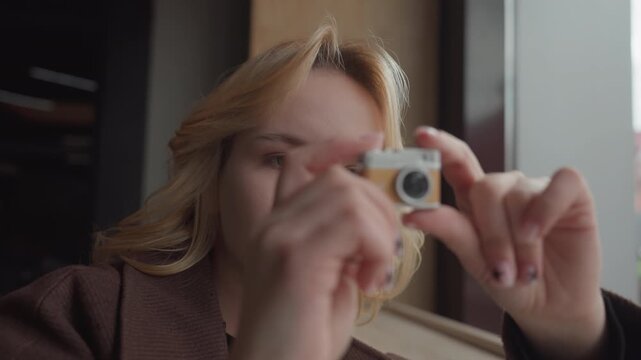 Woman with blonde hair holds old fashioned film camera indoors examining its details closely. Woman wears brown textured top and background shows soft natural light from large windows