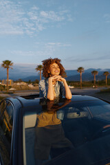 Fototapeta premium Casual woman leaning on car hood, smiling with curly hair, sunny outdoor scene, desert landscape with palm trees, relaxed lifestyle, warm glow, carefree mood, candid moment in bright light.