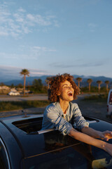Fototapeta premium Young woman leaning out of a car window, laughing with curly brown hair in a denim jacket, sunset light and palm trees in the background, mountains on the horizon.