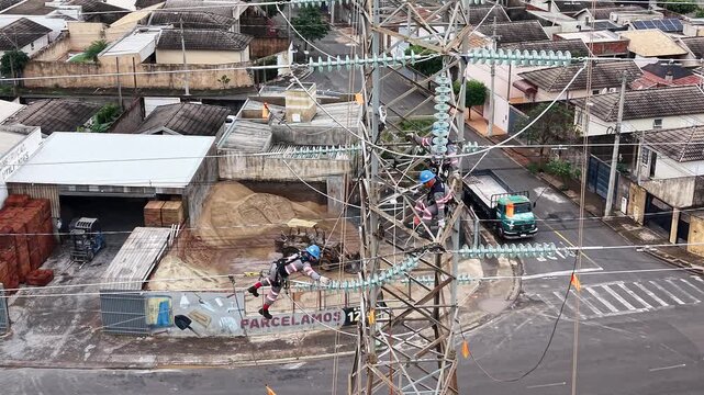 Workers in safety gear perform maintenance on high voltage power lines above a residential street and industrial area