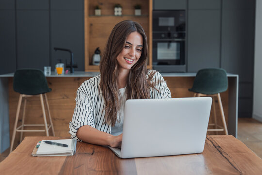 Woman manages home finances on a laptop at her kitchen table, typing beside a notebook. Home finances, budgeting and digital banking support household money management in a bright home office.