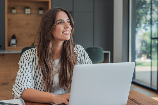 Woman studies online on a laptop at a wooden table, typing while looking away with a thoughtful smile. Home learning, coursework and education fit a bright, quiet study space with natural daylight.