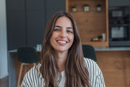 Smiling young woman poses for a portrait in her home kitchen, looking at the camera. Casual indoor lifestyle image communicates beauty, happiness, confidence, modern living and everyday wellbeing.