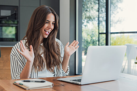Woman greets colleagues during a laptop video call at a wooden table. Her enthusiastic wave and smile support remote work, workplace communication, online meetings and a welcoming team connection.