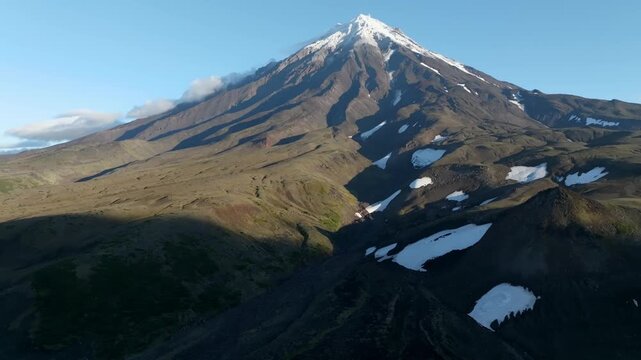 Majestic Koryaksky stratovolcano surrounded by rugged volcanic terrain and snow patches rises into clear blue sky in Kamchatka Krai, Russia, capturing remote wild nature scenery