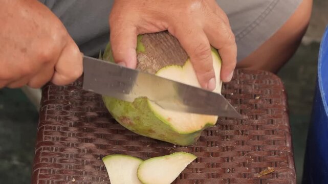A high-angle, close-up shot captures a vendor skillfully shaving the outer husk of a fresh, green young coconut using a large cleaver knife.