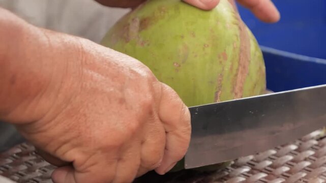 Close-up shot of a man's hands using a sharp cleaver knife to cut open a fresh, young green coconut.