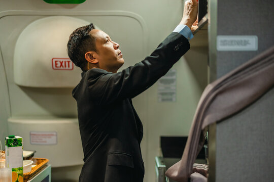 Male flight attendant checking cabin compartment near emergency exit door in airplane