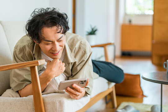 ソファでくつろぎながらスマートフォンを見る笑顔の若い男性。家で過ごすリラックスタイム