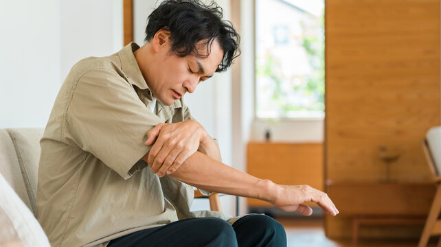 家で肘を押さえて腕の痛みに悩む40代男性。肘関節の痛みや筋肉疲労・体の不調のイメージ