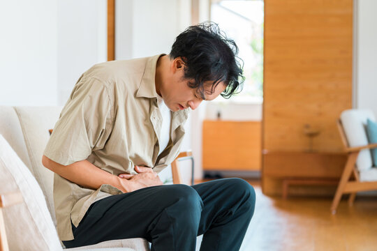 家のソファでお腹を押さえて腹痛に悩む40代男性。胃痛や消化不良・体調不良のイメージ