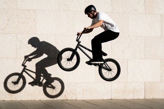 Young male bmx rider performs bunny hop stunt in urban street. Athlete jumps on black bicycle against light stone wall. Large shadow of cyclist projected on building surface. Extreme sport action