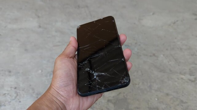 Hand holding a heavily damaged smartphone with a severely cracked and shattered glass screen against a outdoor background.